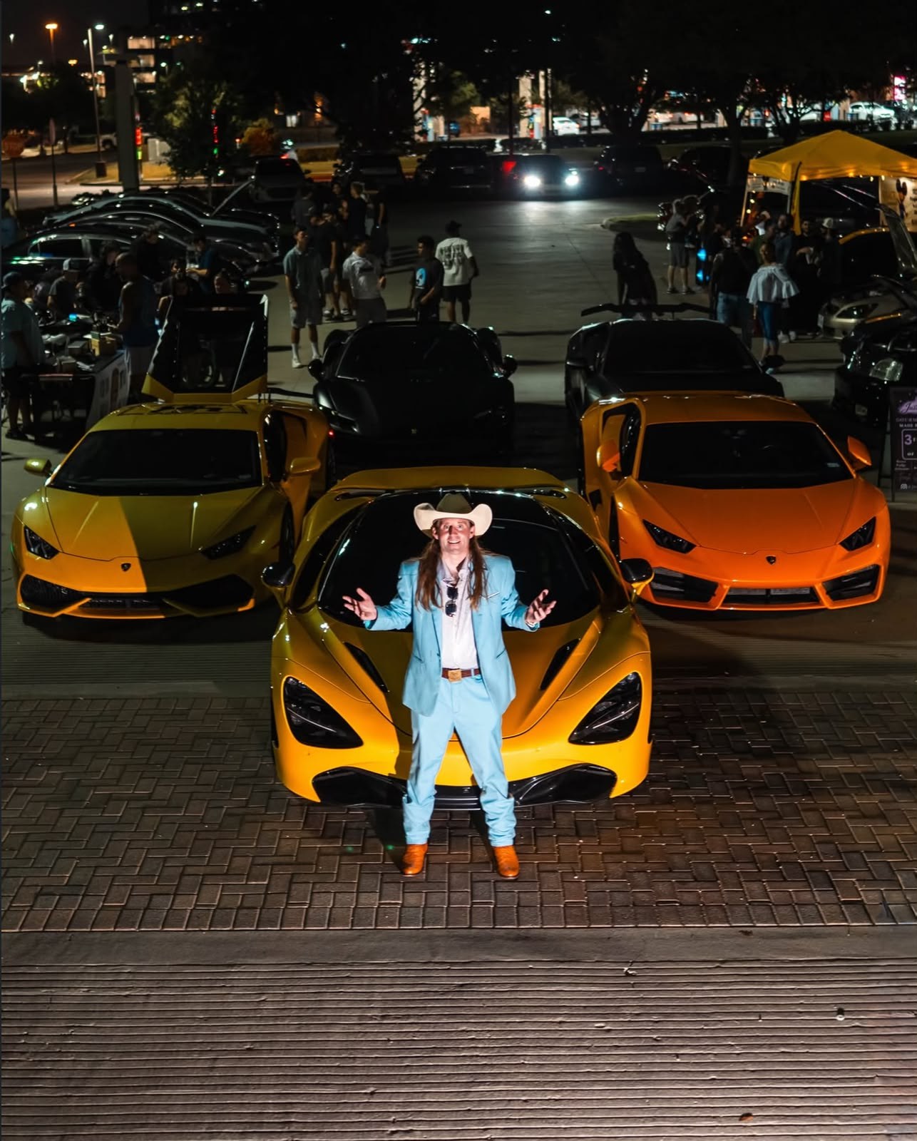 Host in a light blue suit and cowboy hat in front of a yellow McLaren, flanked by two Lamborghinis, with the meet crowd behind